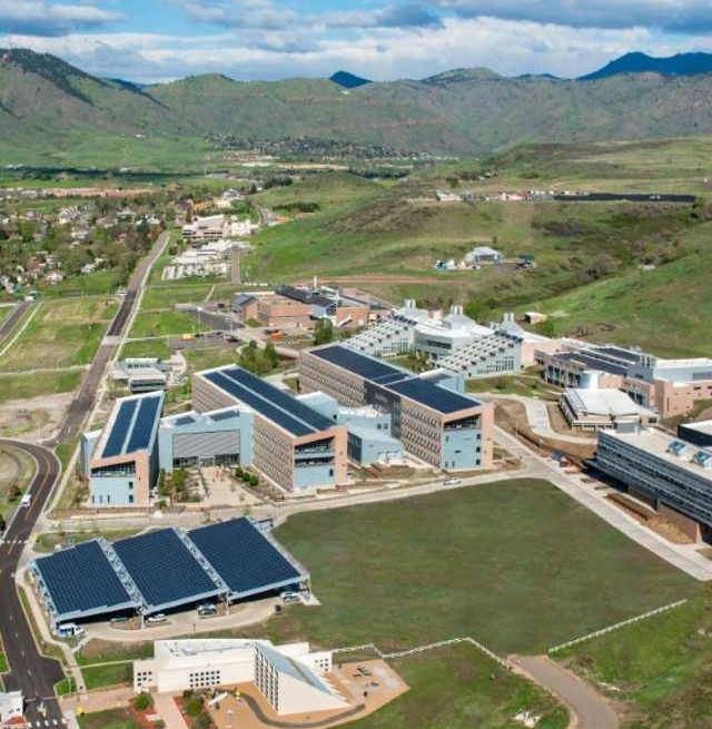 Aerial view of the National Laboratory of the Rockies campus: low
                        research buildings with rooftop solar and solar-covered parking, set
                        against green foothills and mountains under a partly clouded sky.