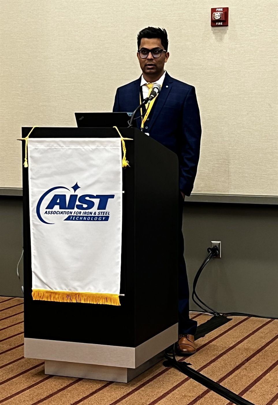 Amogh in a navy suit speaking at a lectern draped with the banner of the
                    Association for Iron & Steel Technology, a laptop open in front of him.