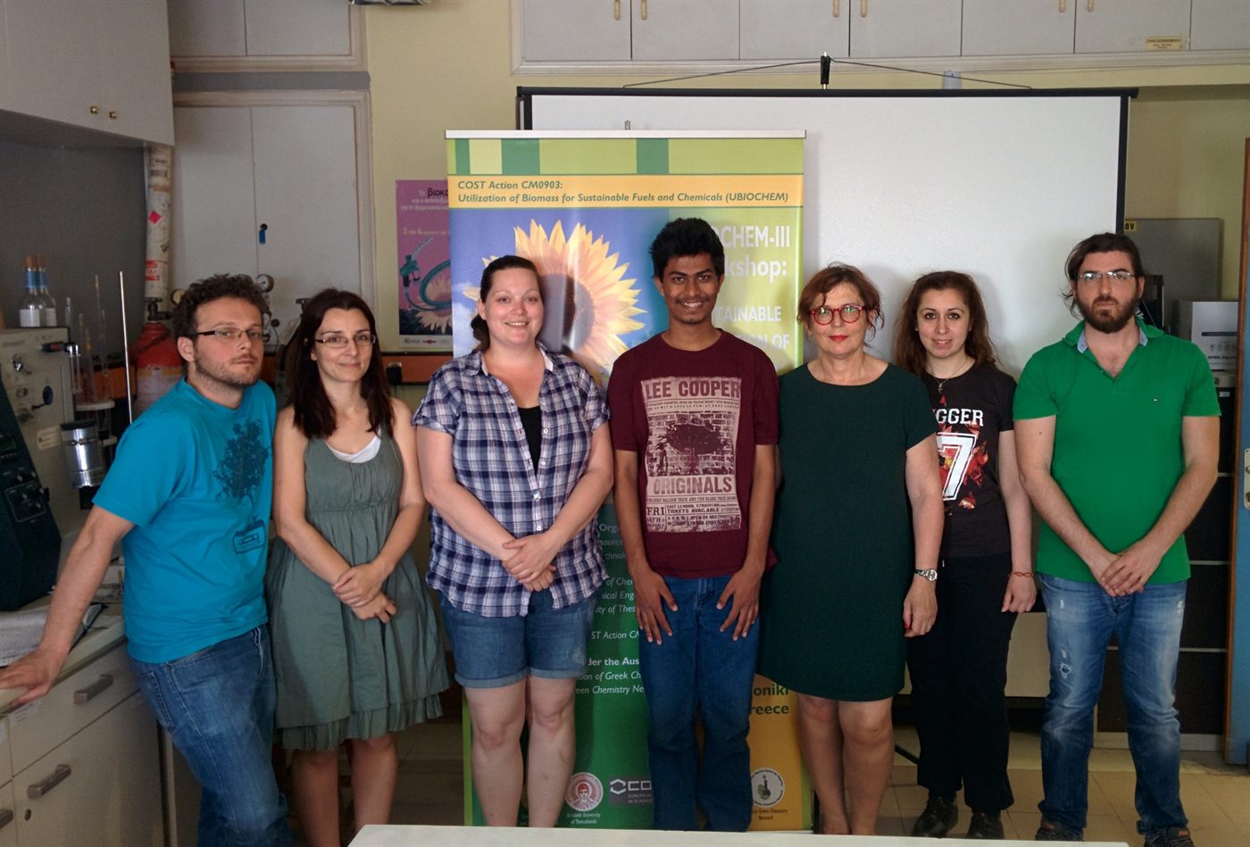 Amogh standing with six lab colleagues in front of a UBIOCHEM banner in a
                    chemical engineering laboratory in Thessaloniki, glassware and a rig
                    visible along the bench behind them.