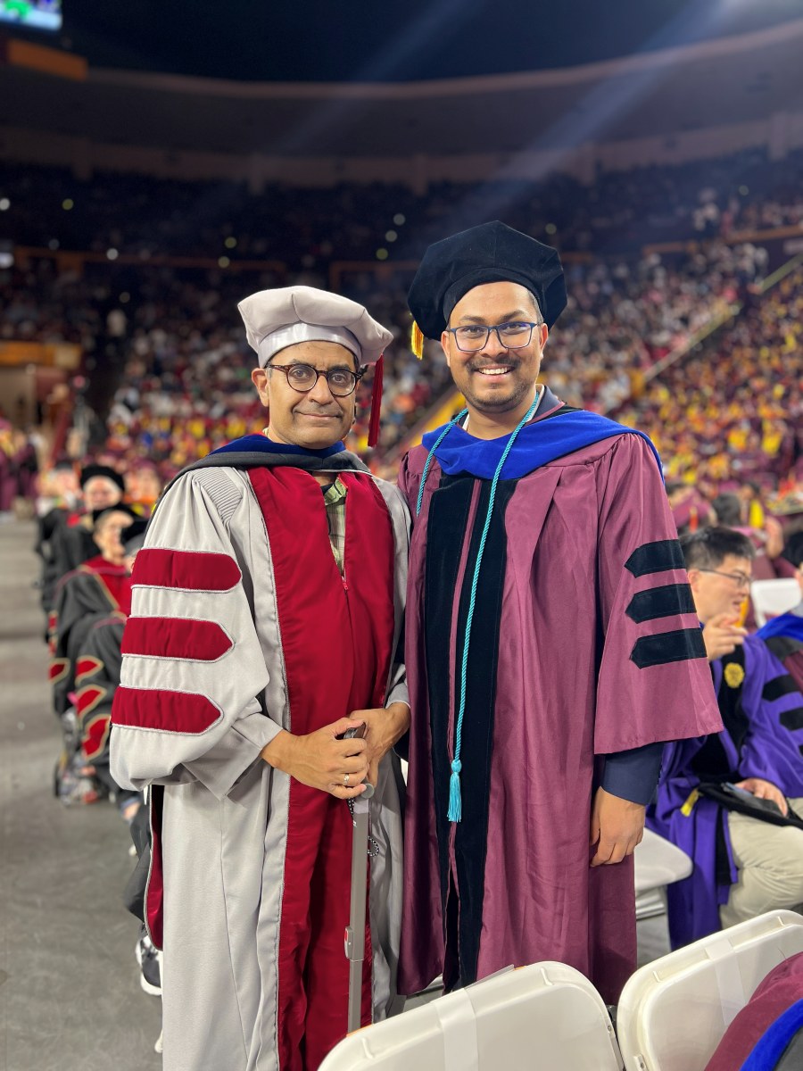 Amogh in maroon doctoral regalia and a black velvet tam, standing beside a
                    faculty member in ceremonial robes in a packed arena at his PhD convocation.