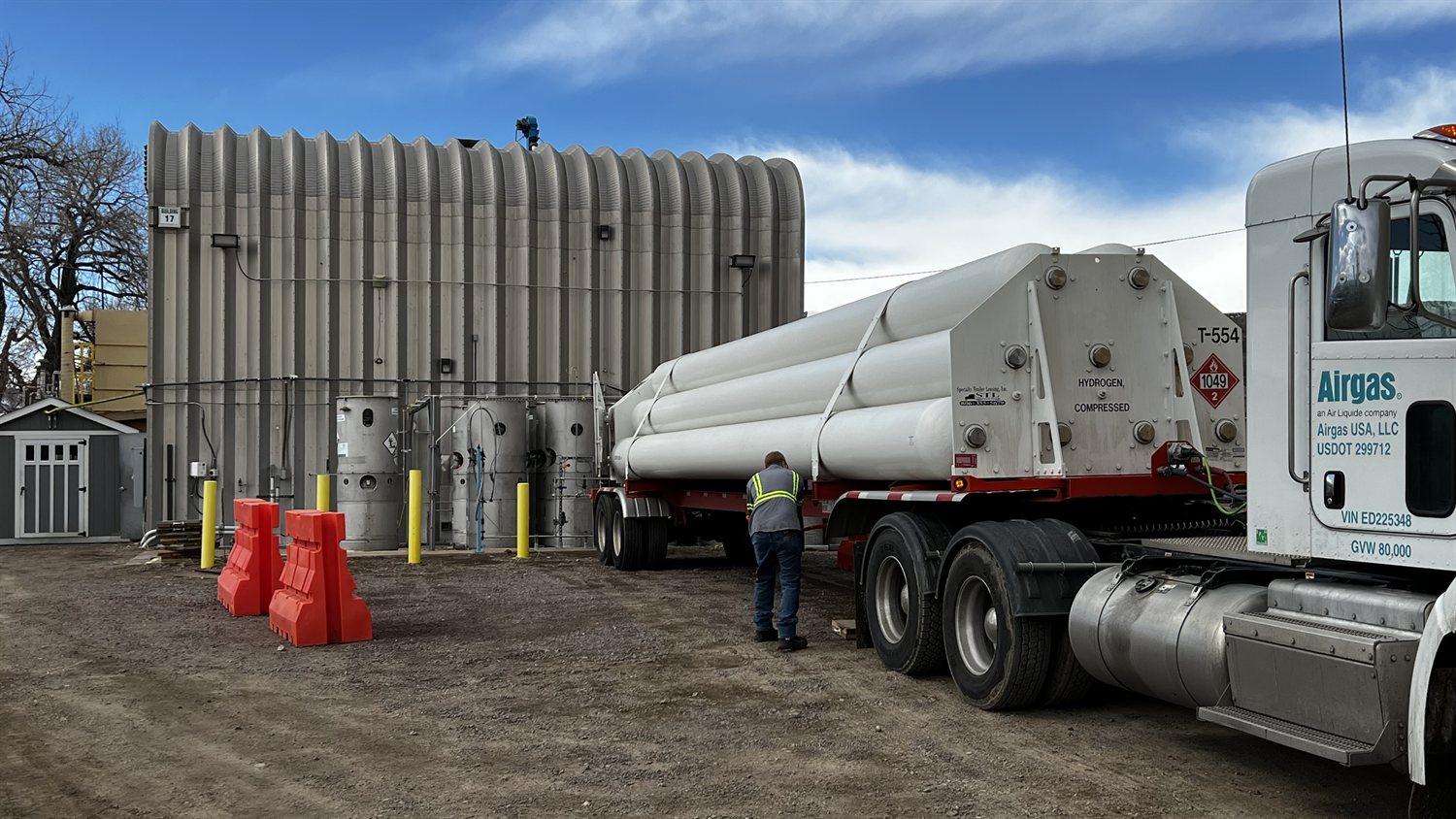 A tanker trailer carrying long cylindrical tubes labelled hydrogen, compressed, parked outside a corrugated metal building, with a worker in a high-visibility vest beside it and small storage vessels behind orange barriers.