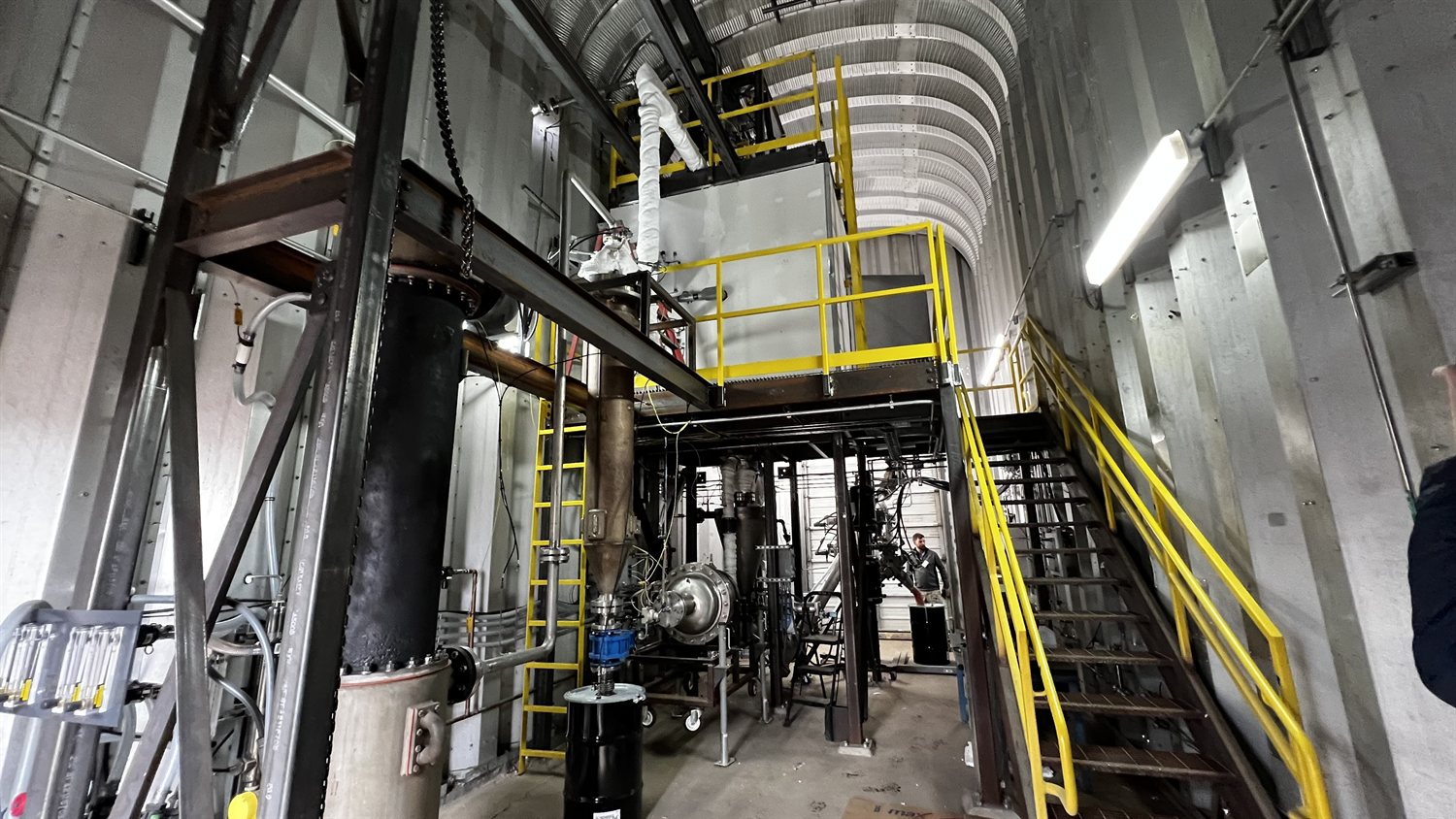 Interior of the pilot plant: a steel structure under an arched corrugated roof, with yellow handrails and stairs, ladders, an insulated vertical vessel and pipework running between platforms.